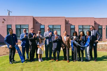 A group of individuals standing outside a building during a ribbon cutting ceremony. A child is holding ceremonial scissors while others are clapping and smiling