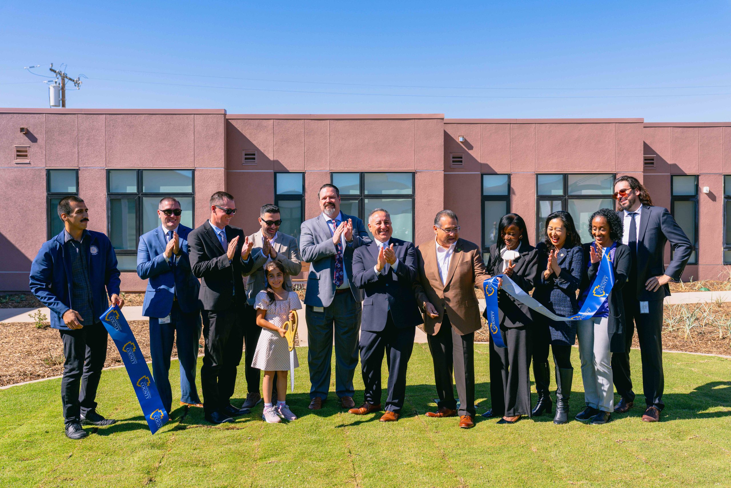 A group of individuals standing outside a building during a ribbon cutting ceremony. A child is holding ceremonial scissors while others are clapping and smiling