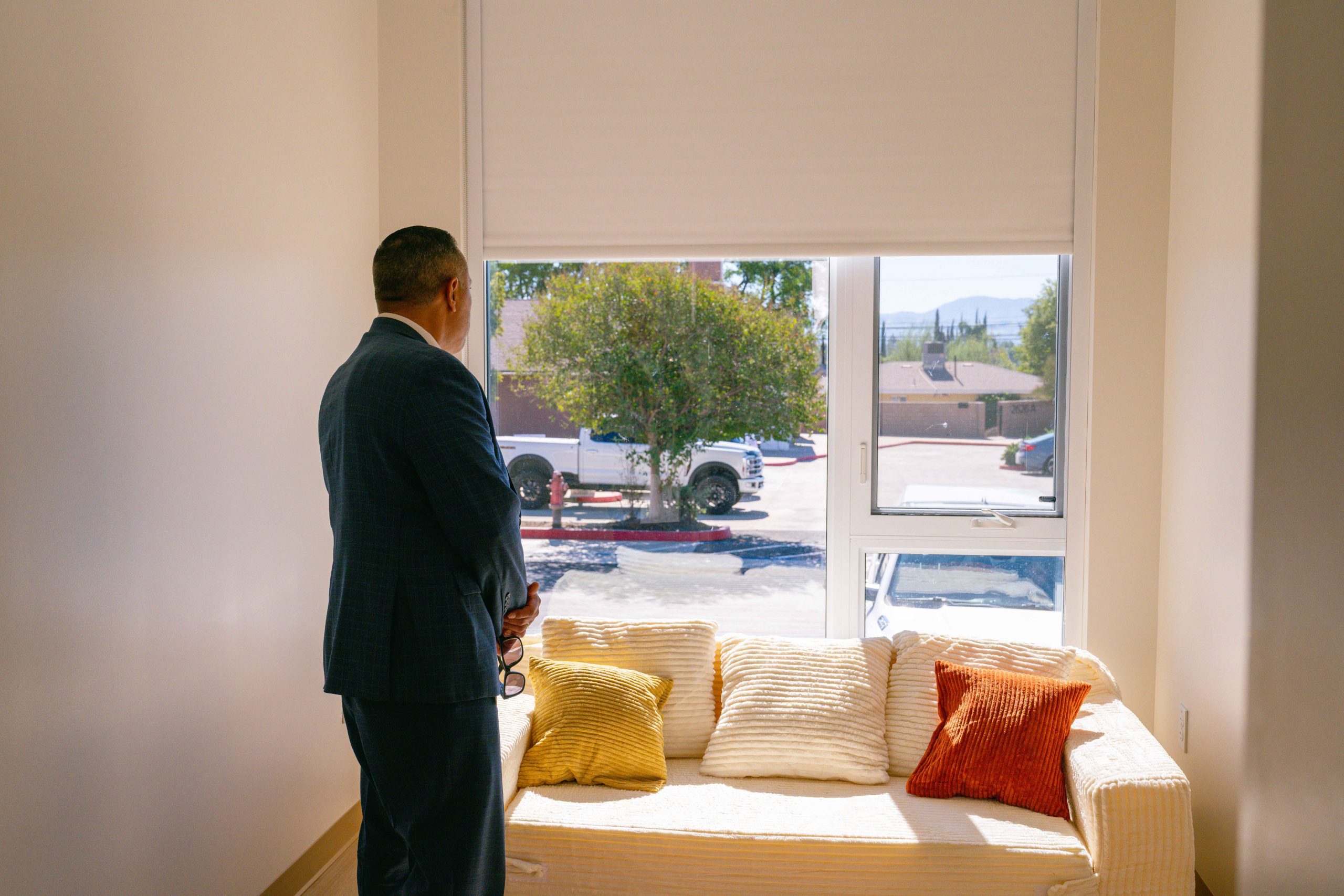 A person in a suit stands in a room, facing a window. Outside, there are trees and a view of a street. A beige couch with a few cushions is positioned near the window.