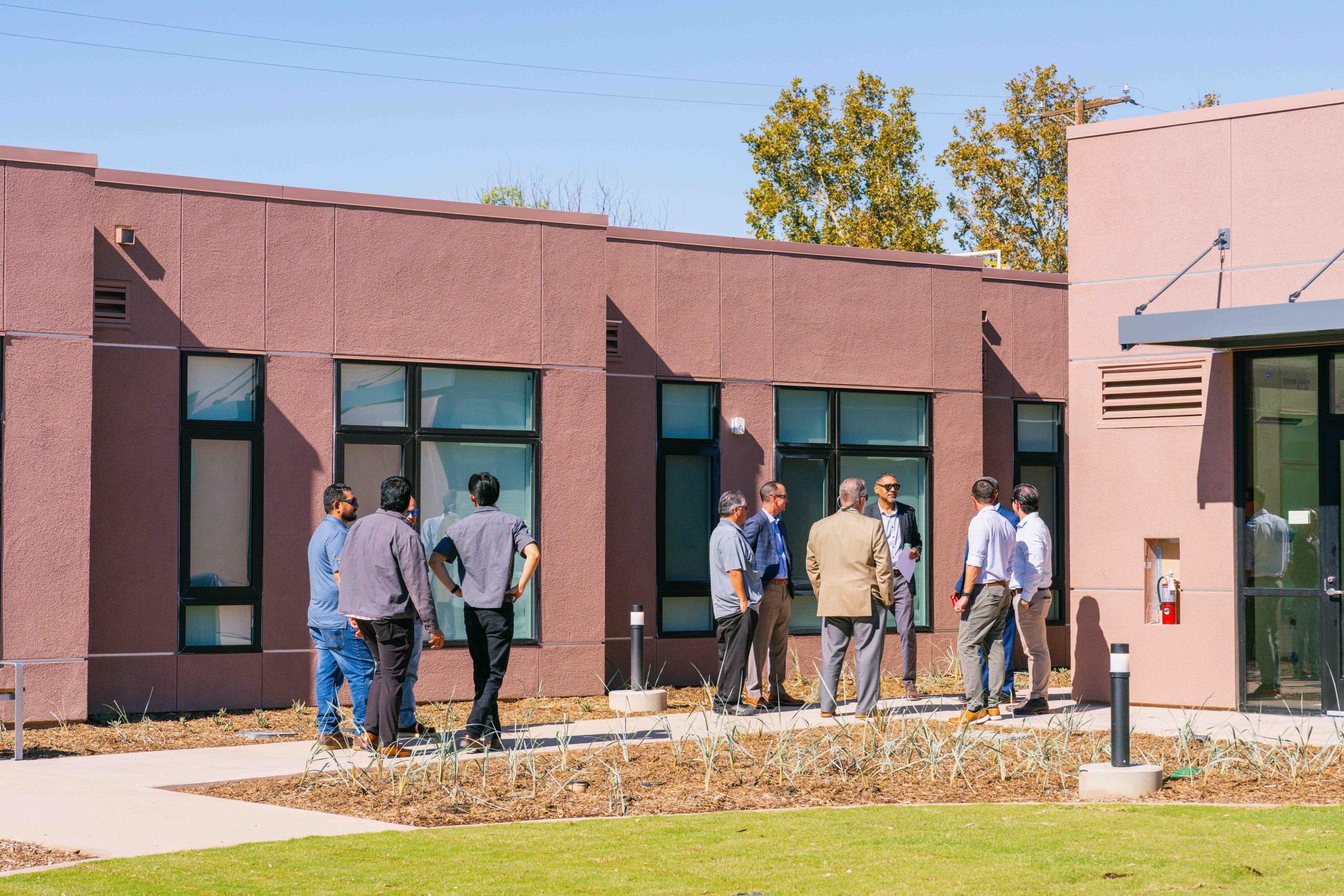 A group of people standing outside a modern, brown building with large windows, having a conversation. The sky is clear, and there are some trees in the background.