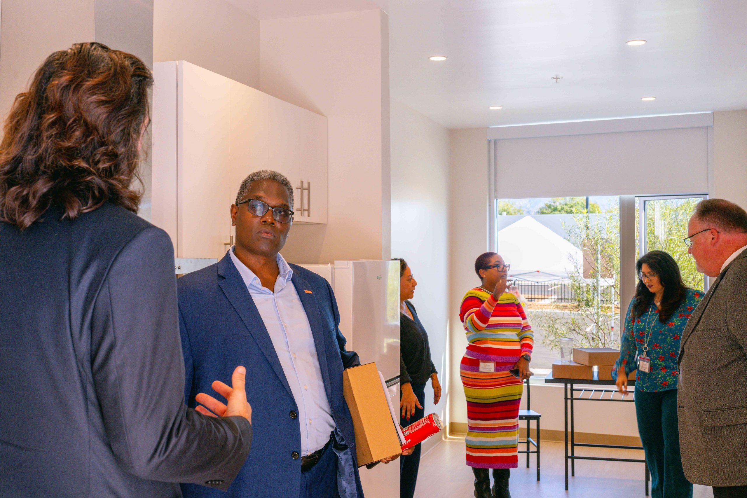 Two people are engaged in conversation in the foreground; one is holding a cardboard box. In the background, three others are talking and standing near a large window. The room has white walls and modern furnishings.