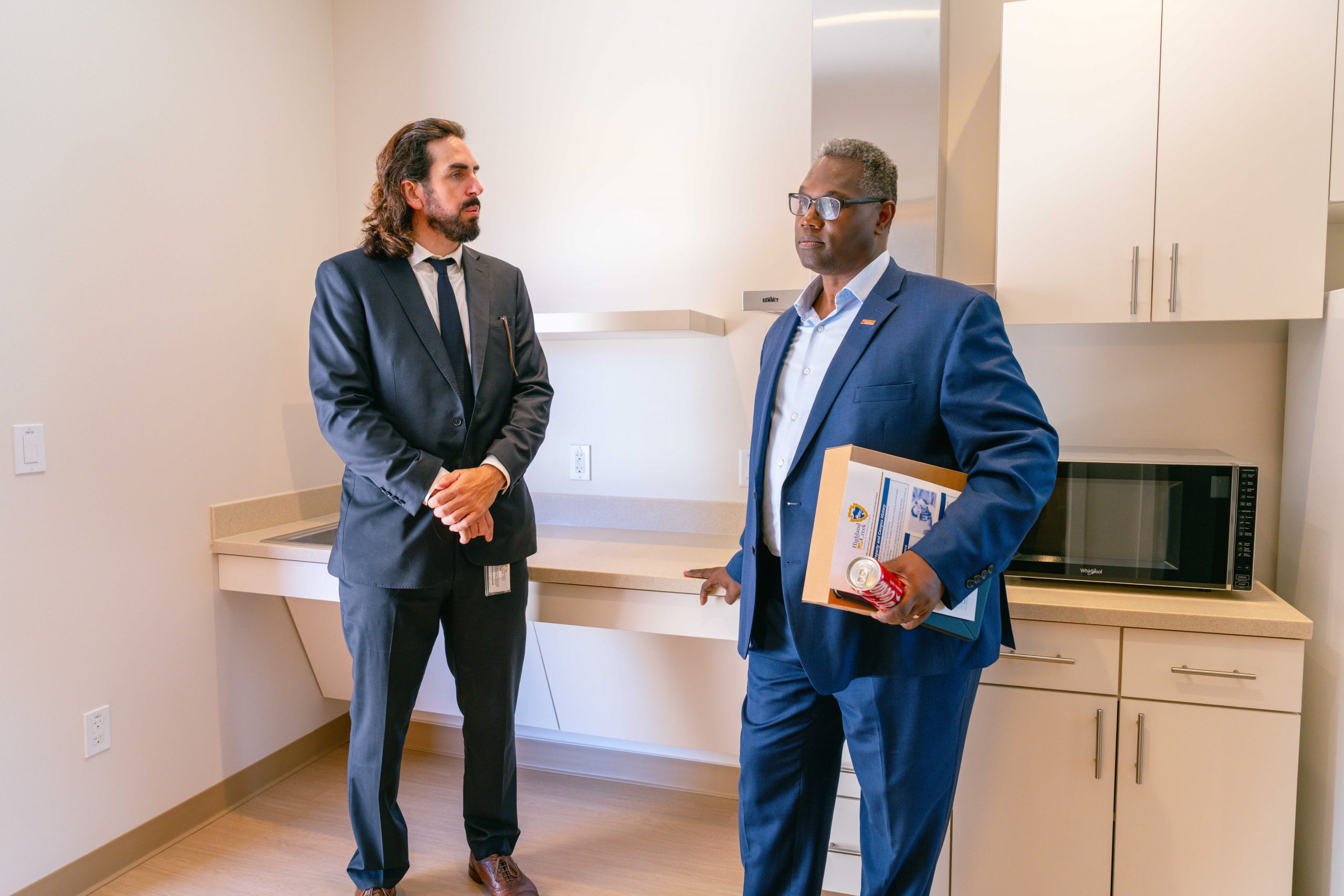 Two individuals in suits are standing and talking in a small office kitchen. One is holding a folder. A microwave is placed on the countertop.