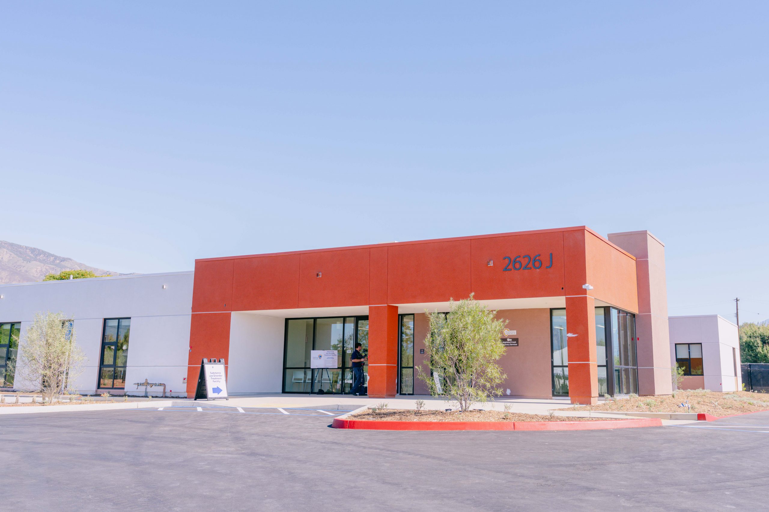 A modern building with a flat roof, featuring white and orange-brown sections, large windows, and a sign displaying "2626 J." There is a small sandwich board near the entrance, and a few young trees in front. The sky is clear.