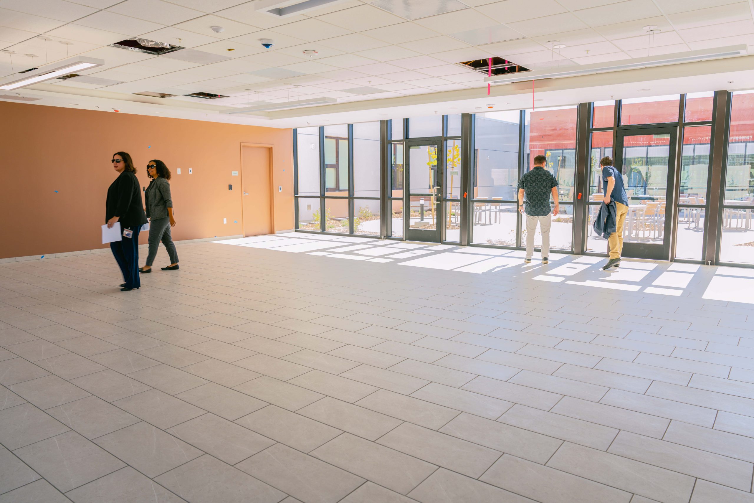 A group of people are standing in a spacious, empty room with large windows allowing natural light to illuminate the tiled floor. The room has a brown accent wall and some ceiling panels are missing. The windows offer a view of an outside area with a red building visible.