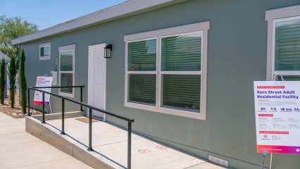 This image shows the exterior of a gray modular building with a ramp leading to the entrance. Two signs near the entrance read "Kern Street Adult Residential Facility" and provide details about the facility. The building has several windows, and the surroundings include a sidewalk and some landscaping.