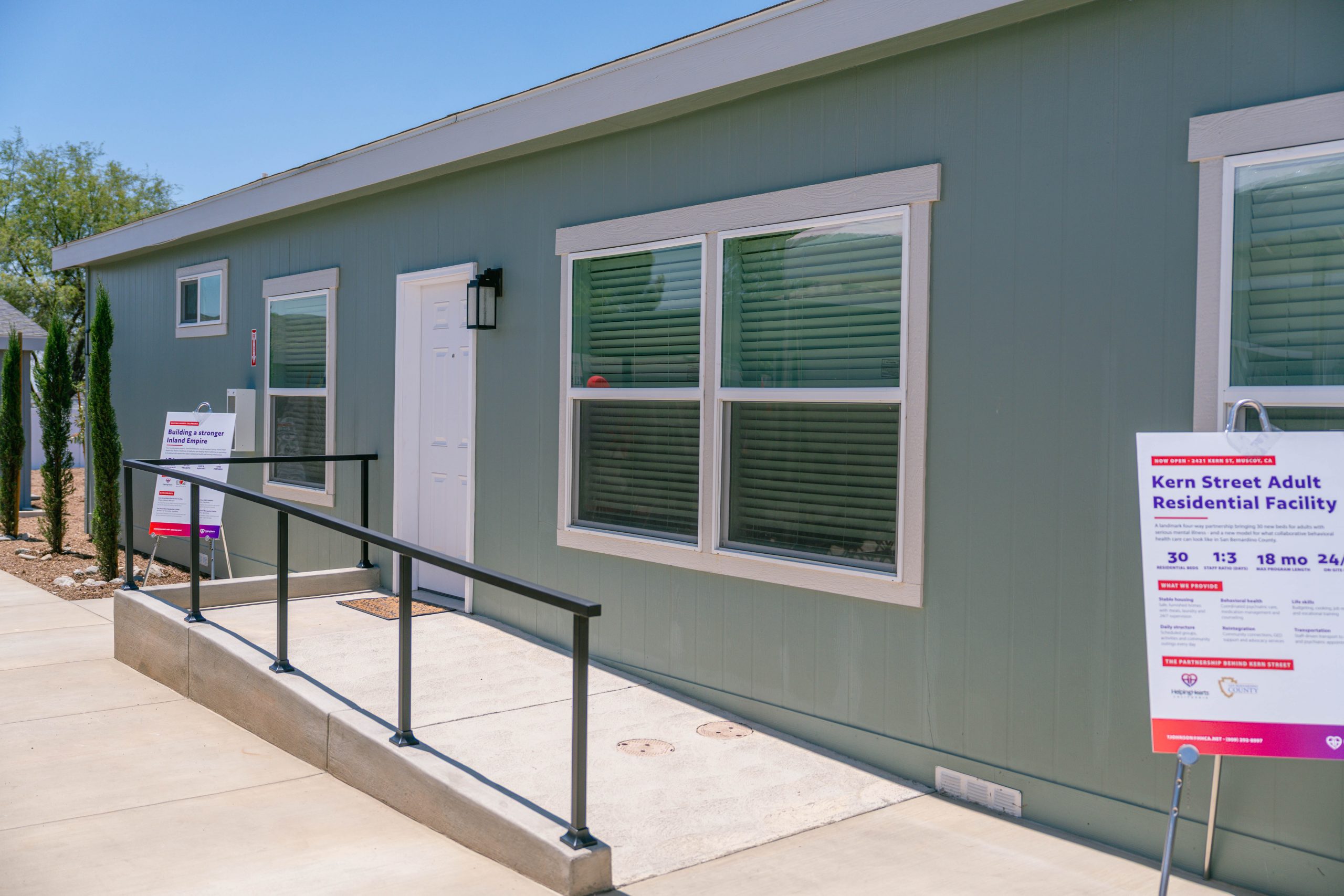 This image shows the exterior of a gray modular building with a ramp leading to the entrance. Two signs near the entrance read "Kern Street Adult Residential Facility" and provide details about the facility. The building has several windows, and the surroundings include a sidewalk and some landscaping.
