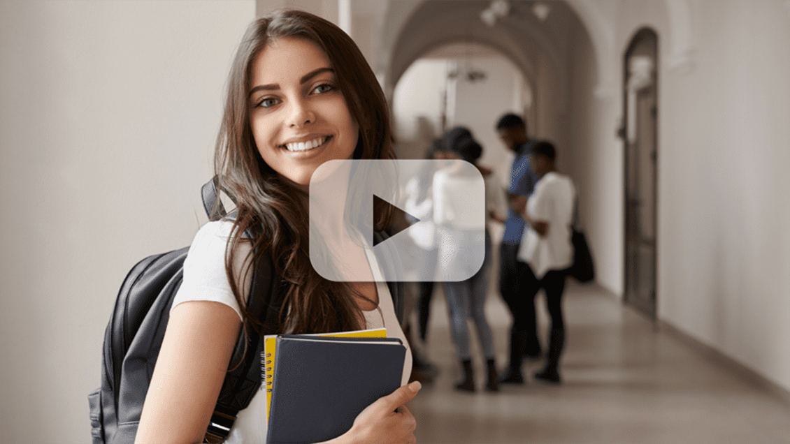A play button lays over an image of a college student in the hallway of an educational institution, smiling and holding notebooks with a gray backpack on. A group of youth stands in the hallway behind her.