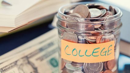 A glass jar labeled "College" is filled with coins. In the background, there are open books and several twenty-dollar bills, suggesting saving money for education.
