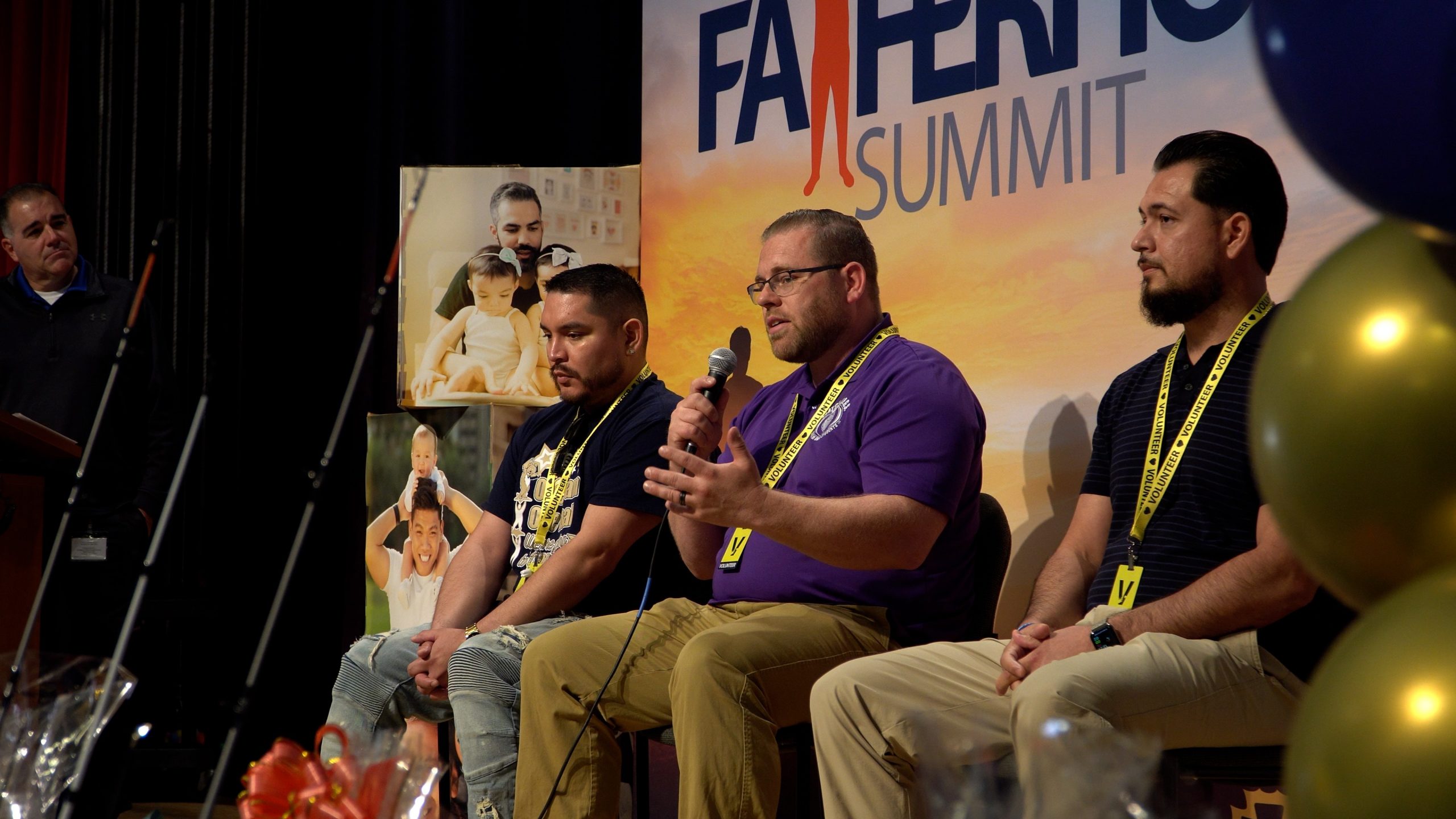 Four individuals seated on stage at the Fatherhood Summit with one holding a microphone and speaking. A banner with event title and images of parents and their children.