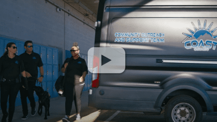 COAST Ontario members in uniform walk past blue lockers beside their team van with a play button overlay, indicating a video.