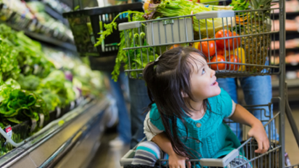 A child sits at the bottom of a shopping cart filled with fresh vegetables at a grocery store.