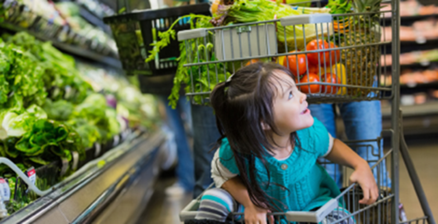 A child sits at the bottom of a shopping cart filled with fresh vegetables at a grocery store.