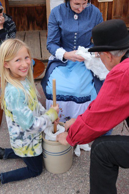 Girl kneels aside a 1800s style milk jar with a stick churning butter aside a woman and man dressed in 1800s period clothing at Calico Ghost Town.