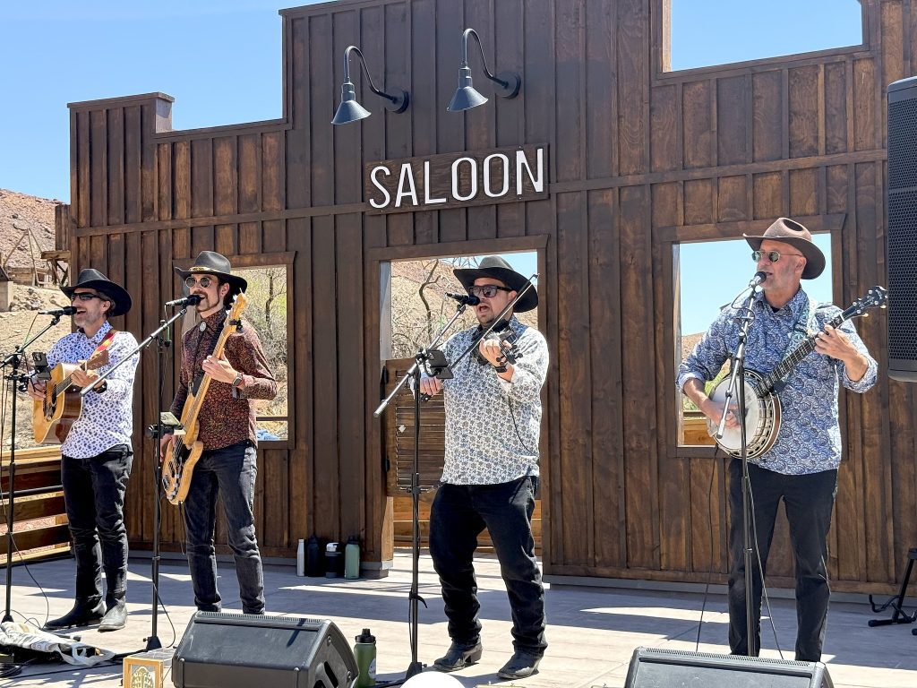 Four men holding guitars stand in front of microphones on a stage in front of a façade designed as an Old West saloon.