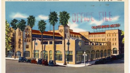 The image shows a vintage postcard of the California Hotel in San Bernardino, California. The hotel features a beige facade, arched windows, and several tall palm trees in front. A few classic cars are parked on the street nearby.