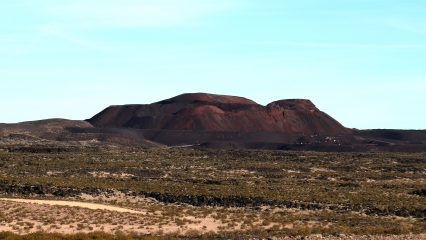 A vast landscape with sparse vegetation stretches towards a large volcanic hill under a clear blue sky.