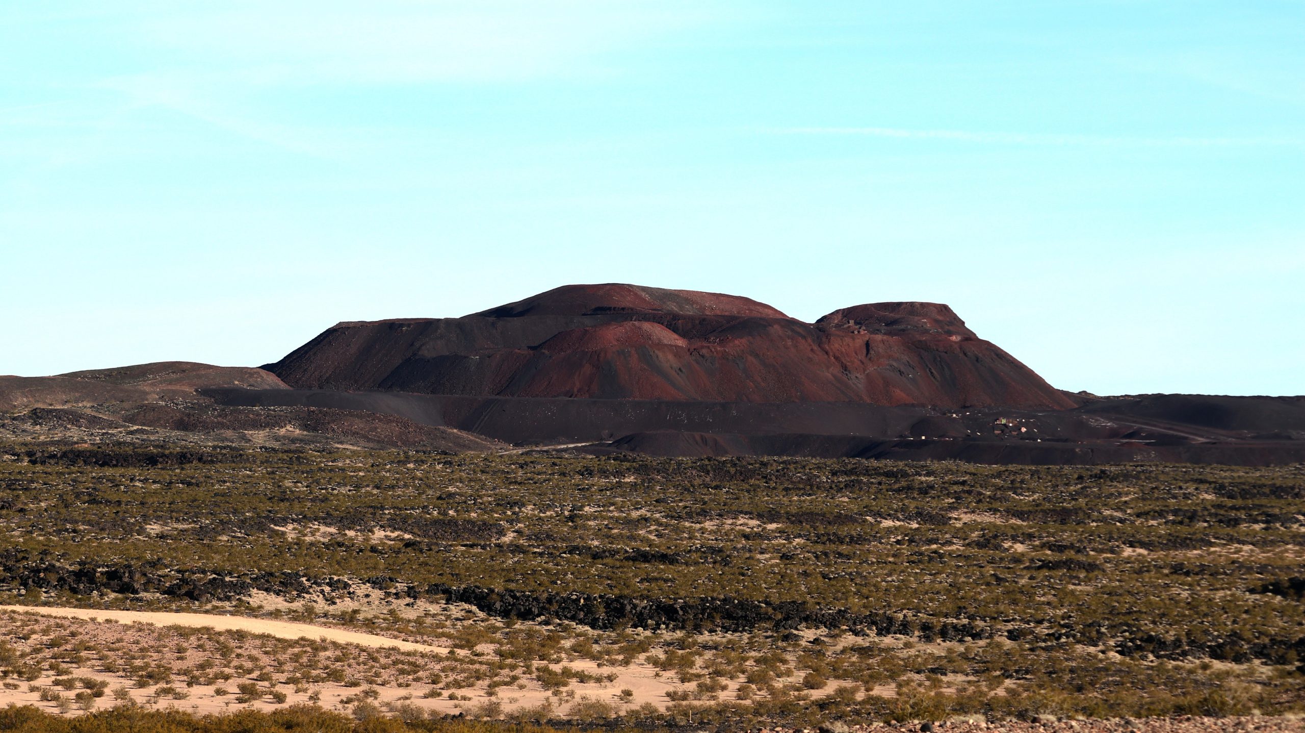 A vast landscape with sparse vegetation stretches towards a large volcanic hill under a clear blue sky.