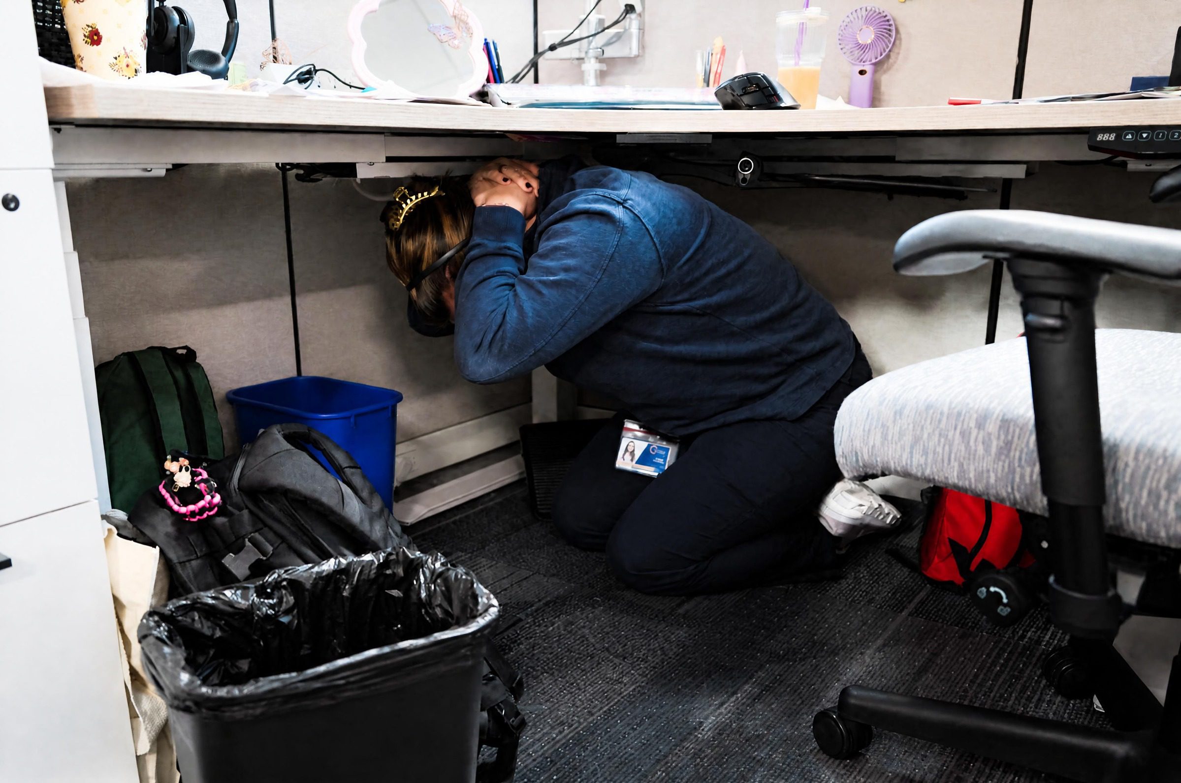 Person crouches under their work desk in a cubicle and covers the back of their neck with their hands.