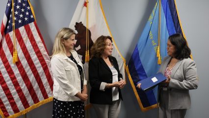 Three individuals are standing in front of several flags. They are engaged in conversation, with one holding a folder.