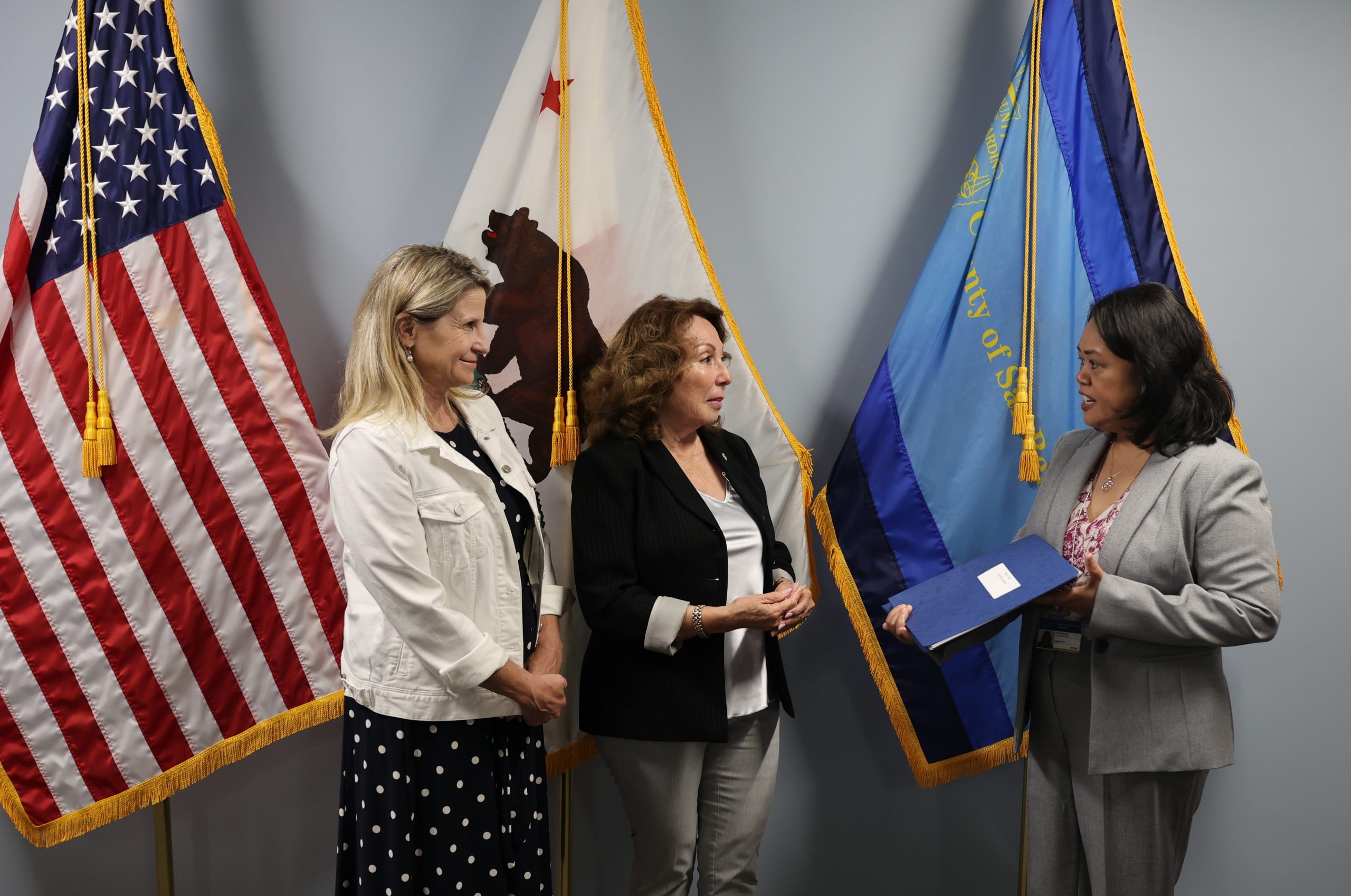 Three individuals are standing in front of several flags. They are engaged in conversation, with one holding a folder.