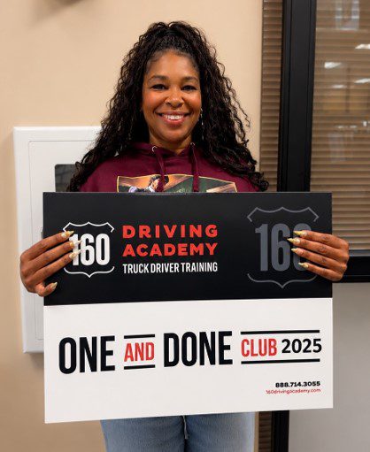 A person stands smiling and holding a sign for "160 Driving Academy Truck Driver Training" that reads "One and Done Club 2025.