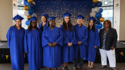 Seven graduates and one faculty member celebrate their achievements against a starry backdrop accented with festive blue and gold balloons.