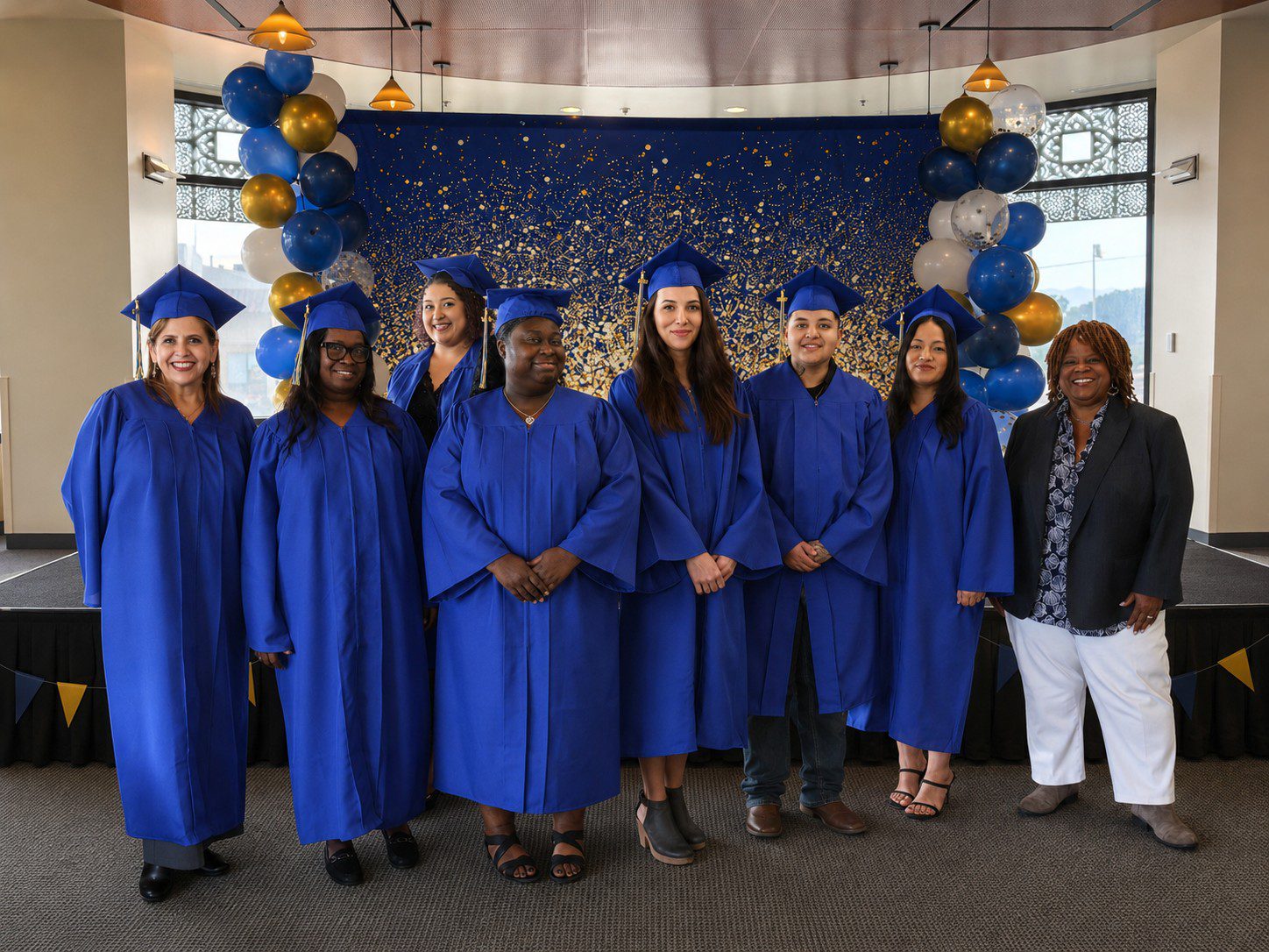 Seven graduates and one faculty member celebrate their achievements against a starry backdrop accented with festive blue and gold balloons.