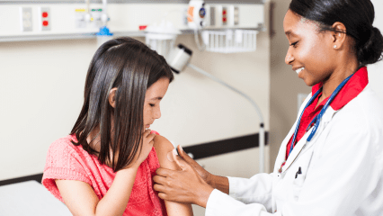 Healthcare provider with a stethoscope carefully puts a bandage on a child’s arm after an injection, as the child watches attentively.
