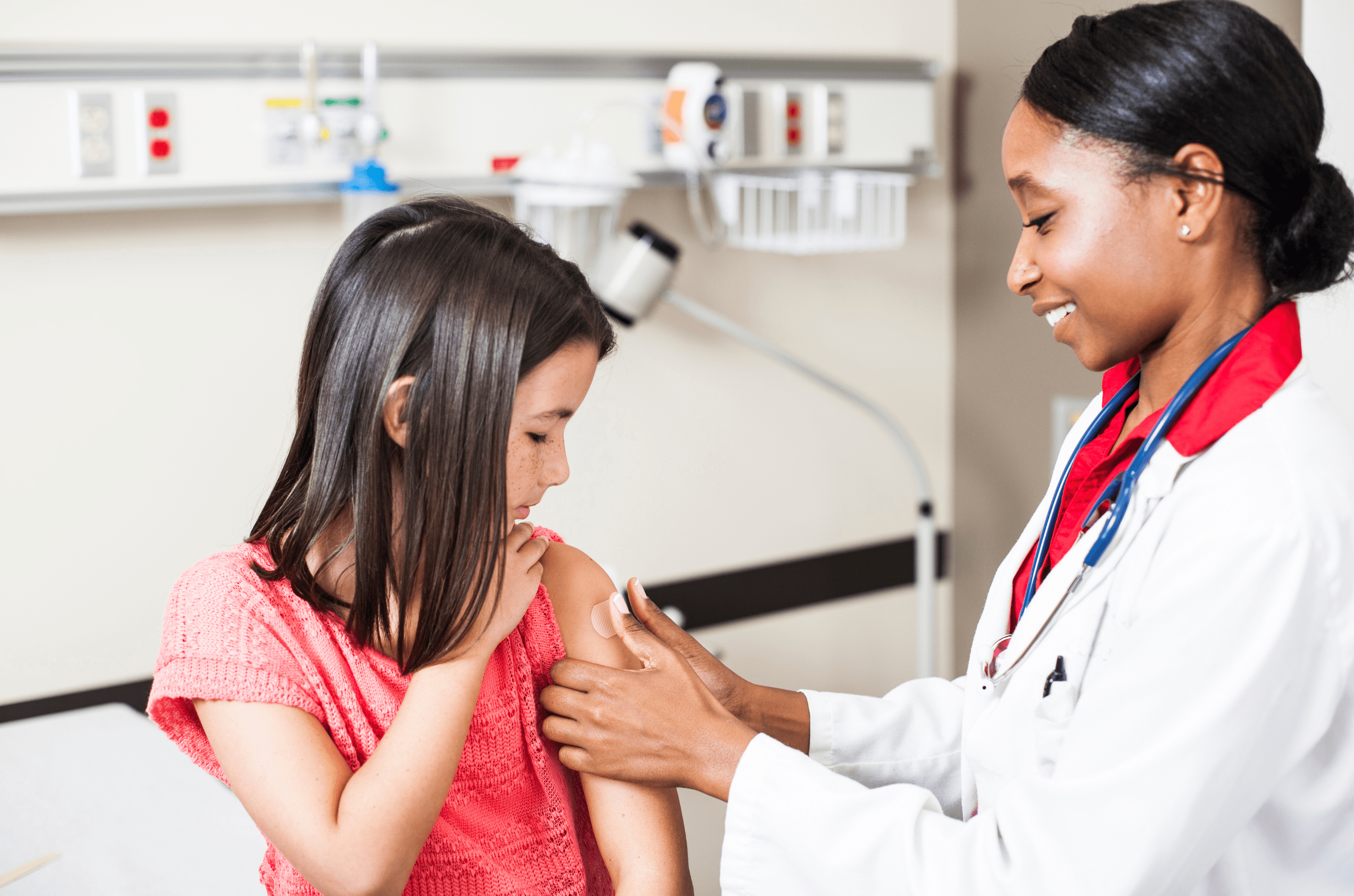 Healthcare provider with a stethoscope carefully puts a bandage on a child’s arm after an injection, as the child watches attentively.