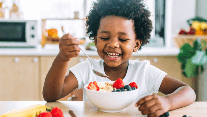 Smiling child eating a healthy bowl of cereal with fruit in a light-filled kitchen.
