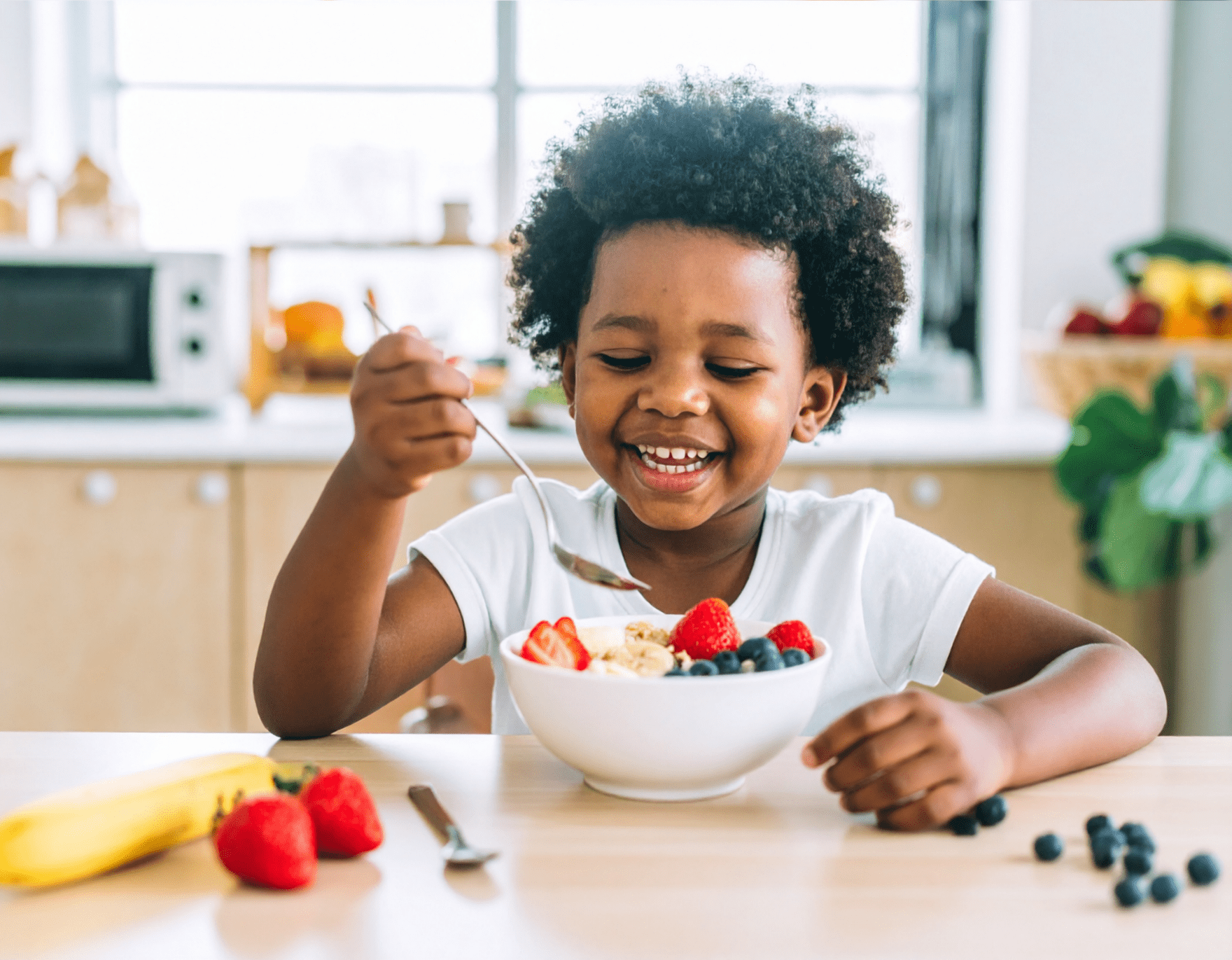 Smiling child eating a healthy bowl of cereal with fruit in a light-filled kitchen.