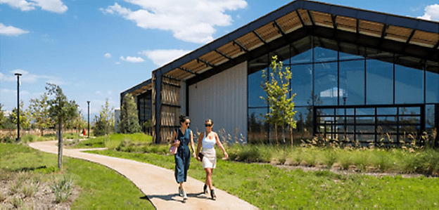 A building with metal, wood framing and glass windows are surrounded by desert landscaping and a cement pathway where two women walk side-by-side.