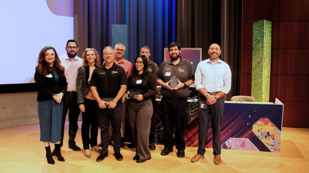Nine individuals standing on stage and smiling at the camera. Two are holding awards.