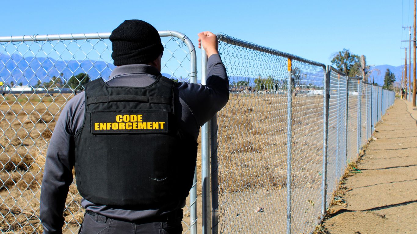 Back view of a code enforcement officer holding onto a chain-link fence around an undeveloped lot.