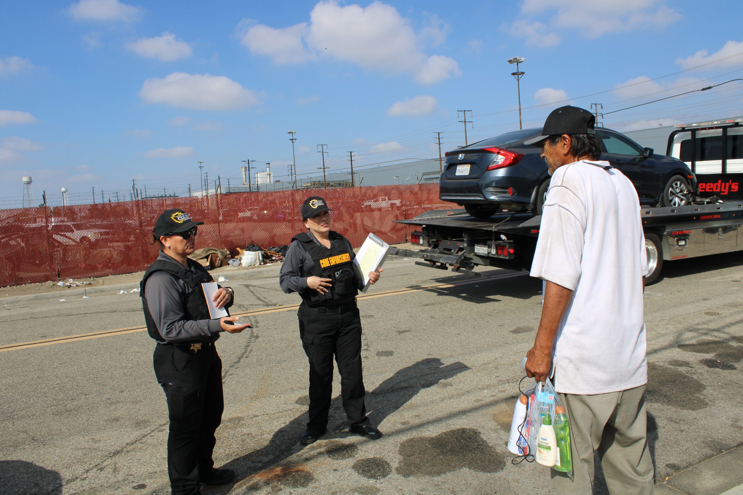 Code Enforcement Operation Photo 3 A street view of two San Bernardino County Code Enforcement Officers talking to a man with a car on a tow bed in the background.