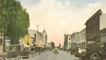 Image depicting old fashioned cars and various commercial buildings down a street.