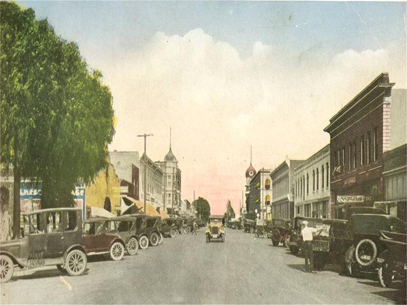 Image depicting old fashioned cars and various commercial buildings down a street.