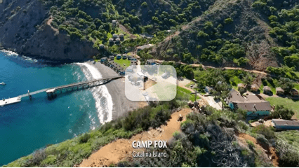 An aerial image of Camp Fox on Catalina Island, part of the Youth Engagement and Enrichment Program, showing a beachside camp, a dock, and forested hills with a play button overlay, indicating a video.