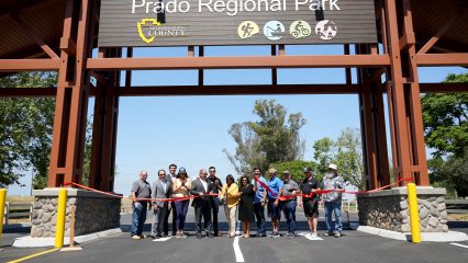 People stand behind a red ribbon under a large Prado Regional Park sign with the County logo and hiking, fishing, bicycling and camping icons.