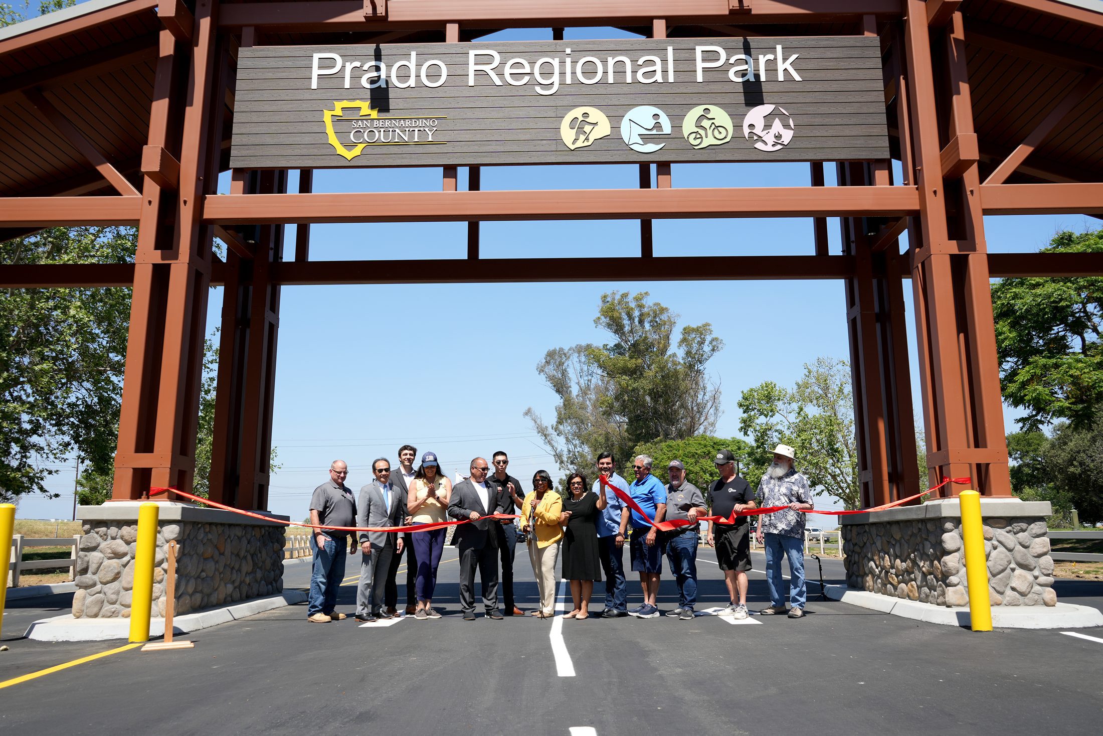People stand behind a red ribbon under a large Prado Regional Park sign with the County logo and hiking, fishing, bicycling and camping icons.