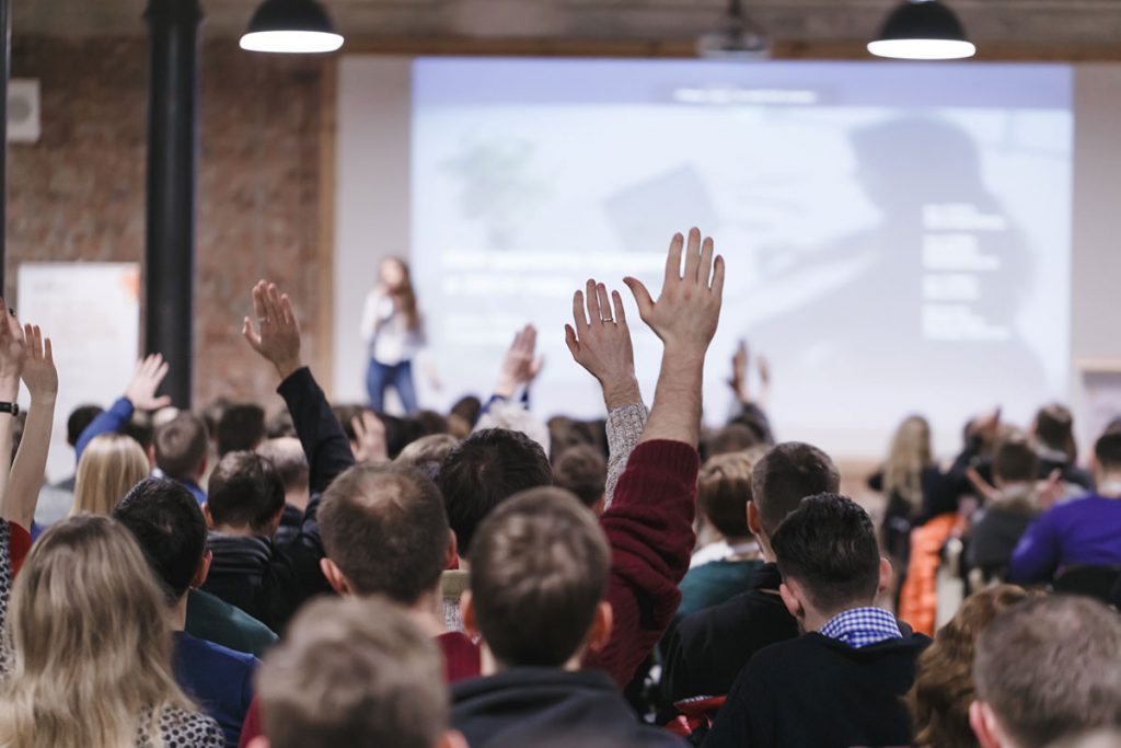 Audience at large meeting, some with hands up