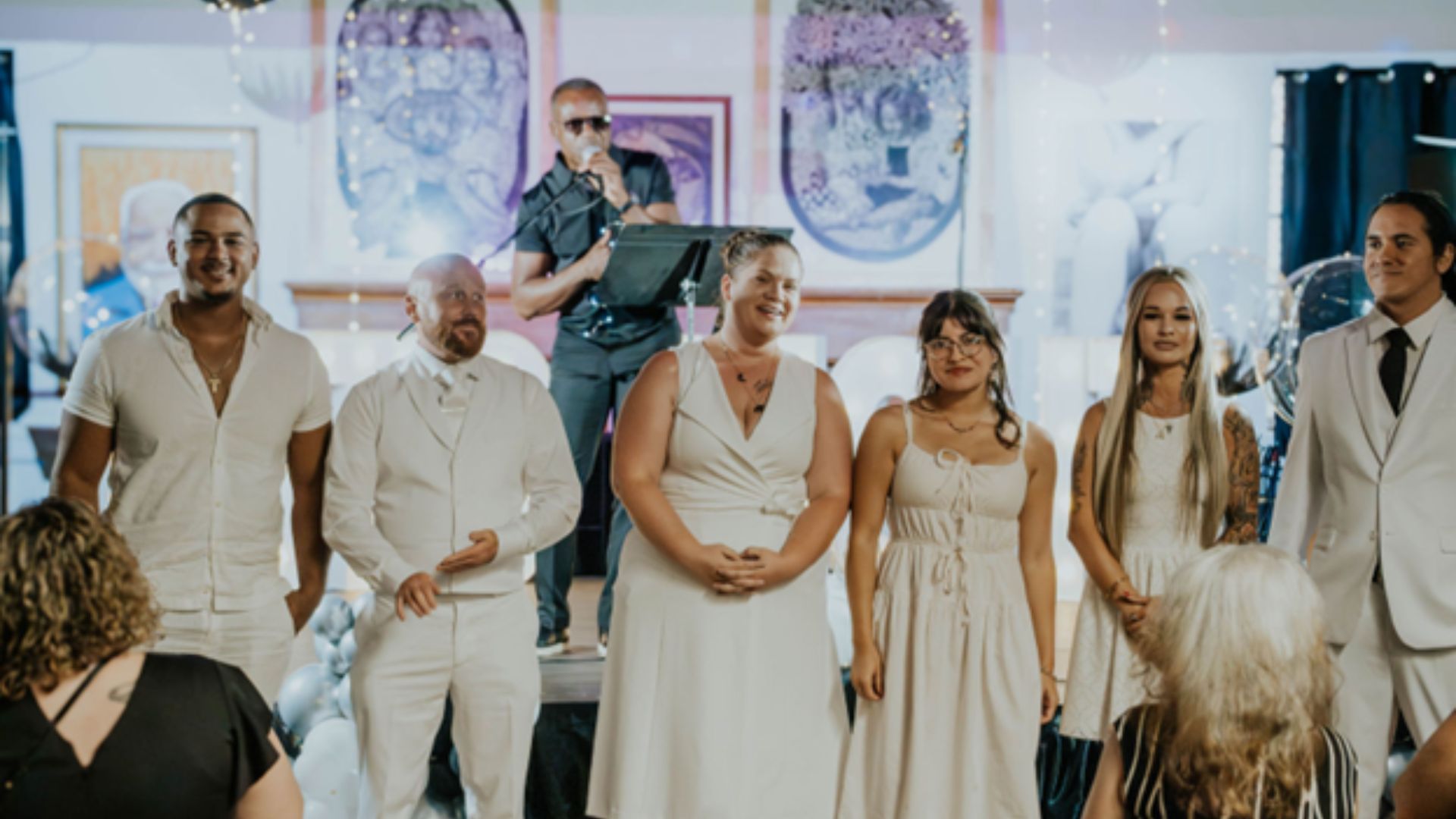 Six people stands at the front of a room decorated for an event, with onlookers seated around them and balloons in the background. An artist performs in the background. Everyone is dressed in formal white attire.