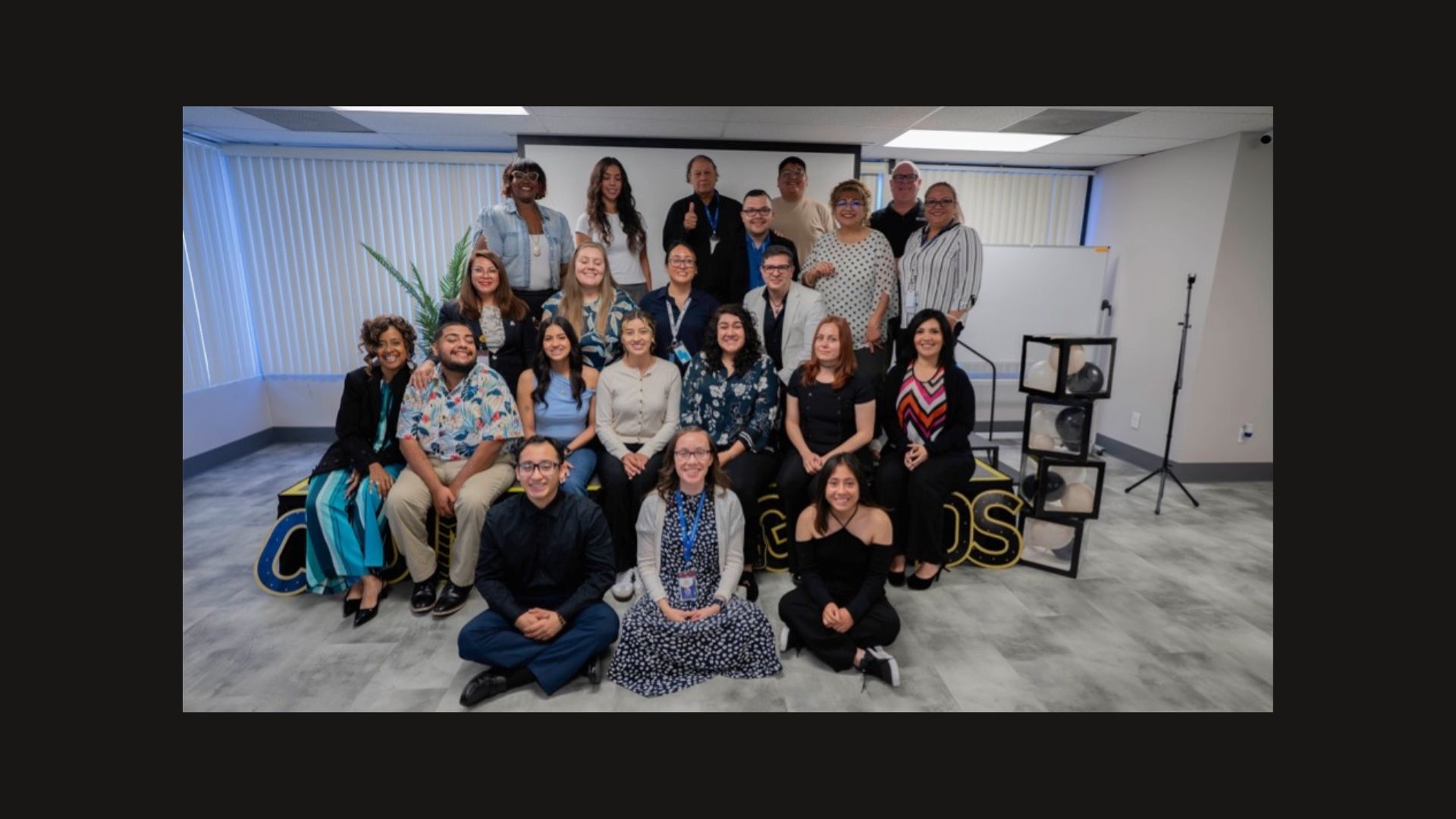 Group of people smiling for a photo in an office environment, standing and sitting in three rows, with a sign reading "Grads” in the background.