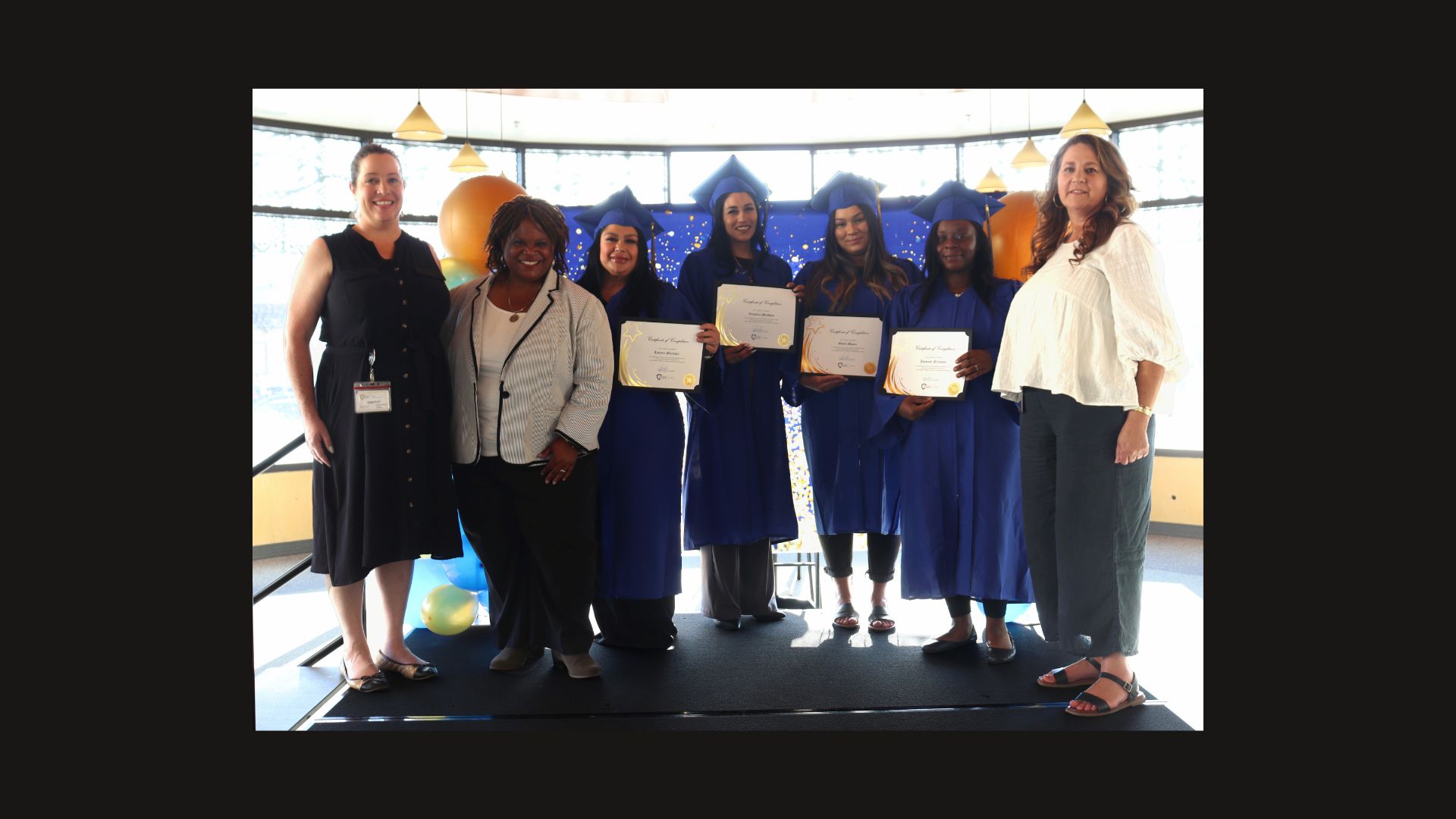 Group of graduates in blue gowns holding certificates, standing with two women on each side, smiling in front of balloons and a decorated backdrop.
