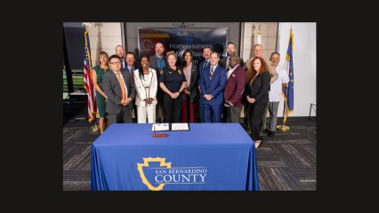 Group of county officials standing behind a table with a sign reading San Bernardino County.