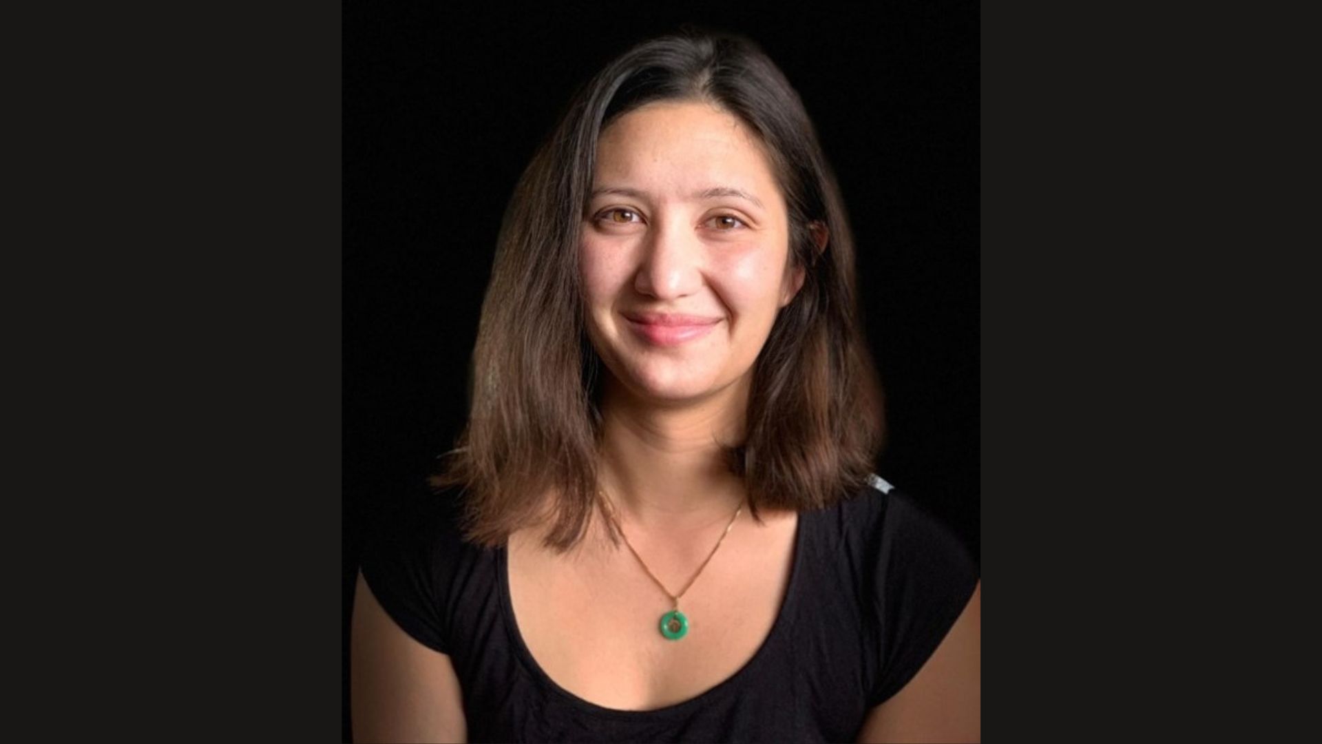 Portrait of a smiling person wearing a black top and a green pendant necklace set against a dark background.