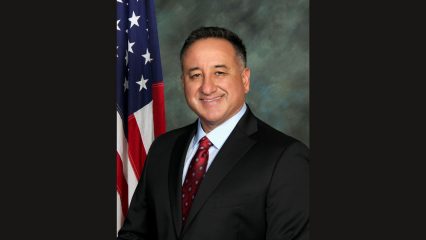 A formal headshot of Supervisor Joe Baca, Jr. in a black suit and red tie with an American flag to his side.