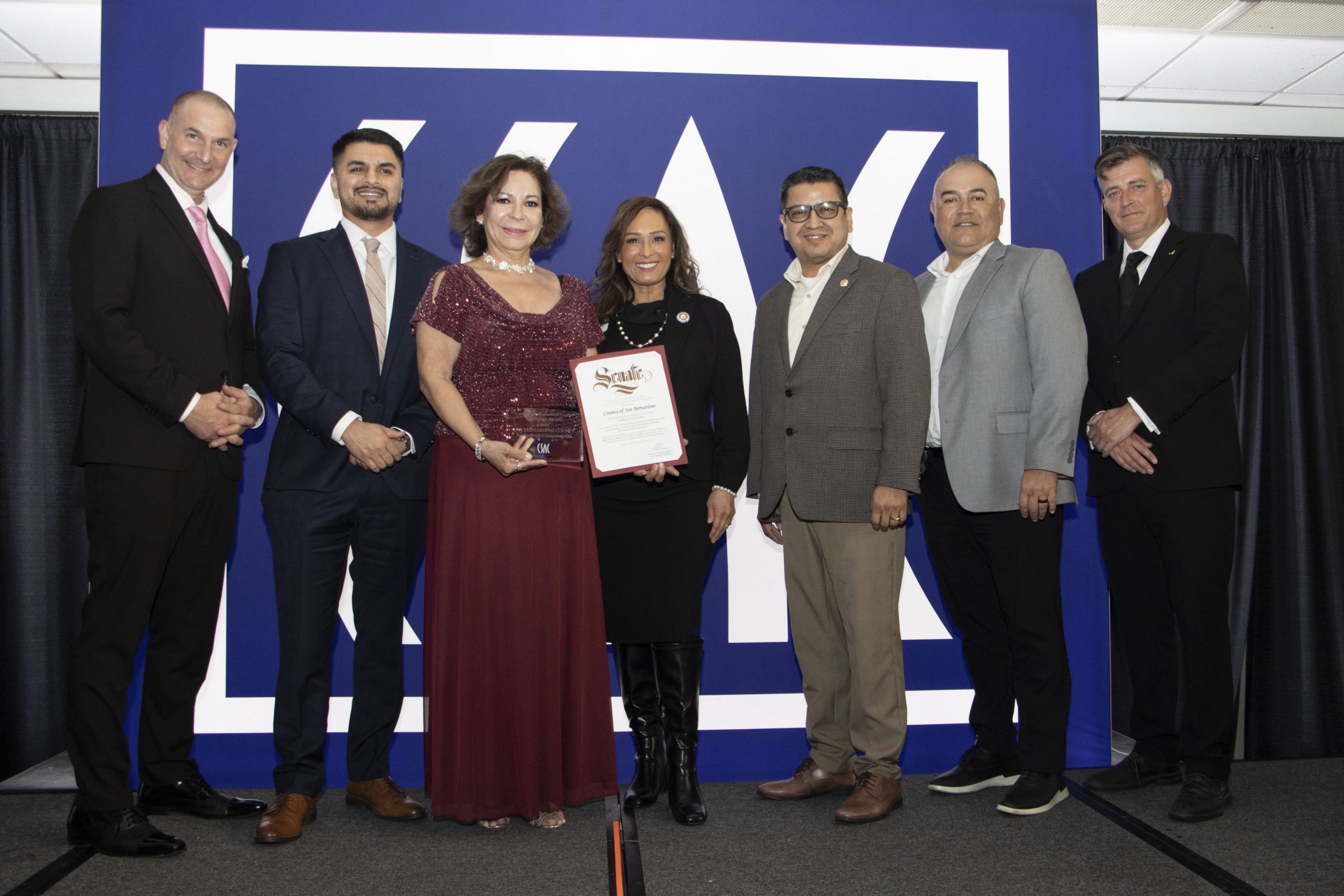 Several officials pose with a CSAC award and State Senate certificate for San Bernardino County’s Emprendedor@s program, smiling in front of a CSAC backdrop.