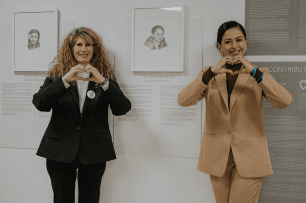 Two women stand in front of portraits, smiling and forming heart shapes with their hands.