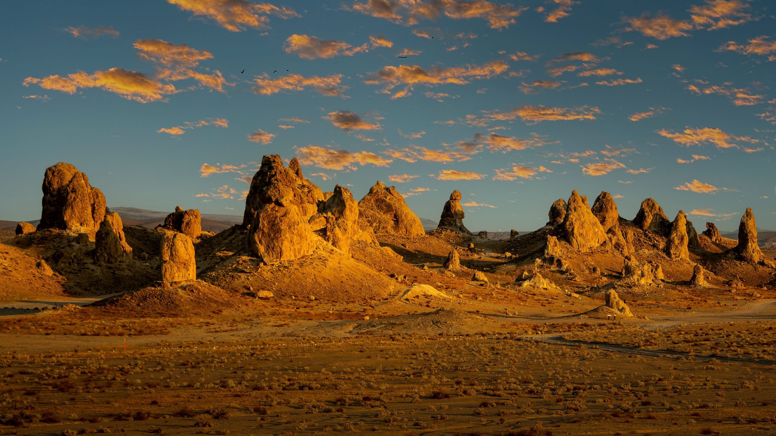 View of the Trona Pinnacles at sunset with golden lighting illuminating the tufa formations under a partly cloudy sky.